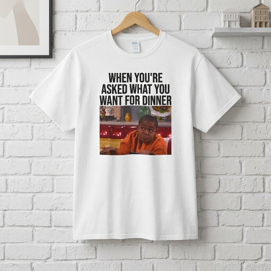 A white t-shirt with a graphic image of a boy shrugging and text above it stating 'When You're Asked What You Want For Dinner' on a hanger against a white brick wall.