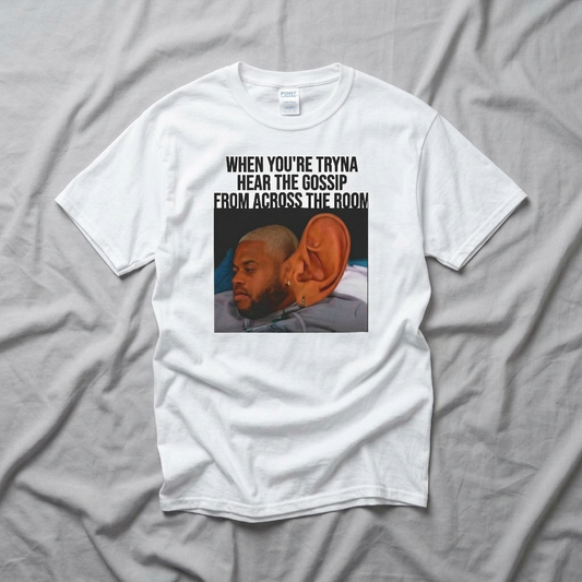 A white t-shirt with graphic design of a man with a large ear and text above it stating 'When You're Tryna Hear The Gossip From Across The Room' on a gray fabric background