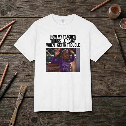 A white t-shirt with a graphic containing a girl with her hands up and text above the image stating 'How My Teacher Thinks I'll React When I Get In Trouble' on a wooden surface with stationery items.