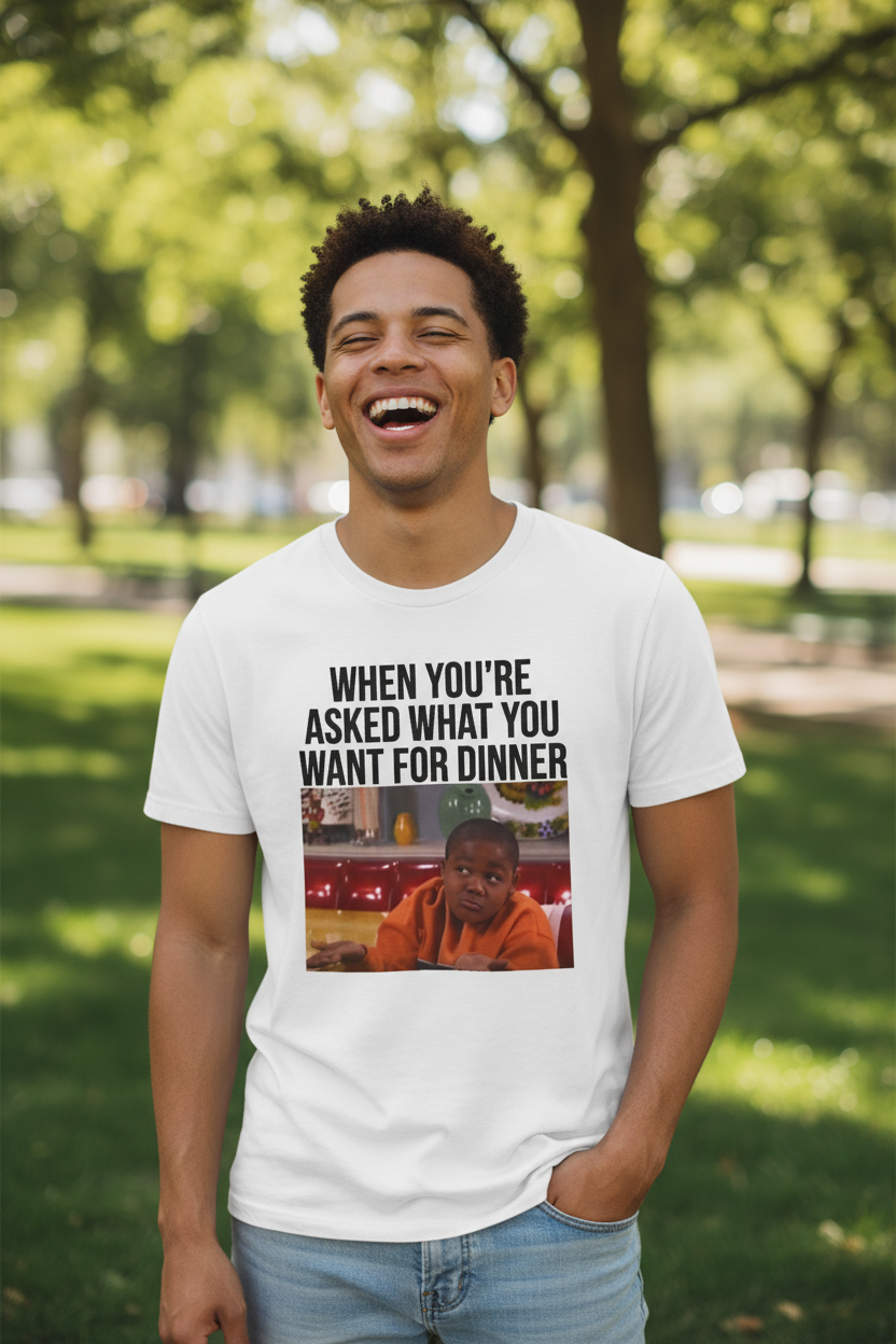A man wearing a white t-shirt with a graphic image of a boy shrugging and text above it stating 'When You're Asked What You Want For Dinner' in a park