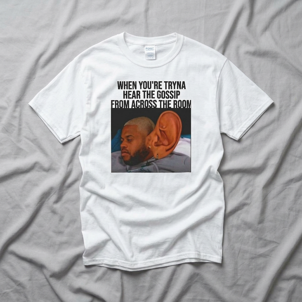 A white t-shirt with graphic design of a man with a large ear and text above it stating 'When You're Tryna Hear The Gossip From Across The Room' on a gray fabric background