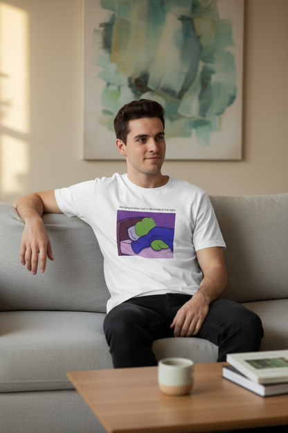 Man sitting on a couch wearing a White t-shirt with a graphic of a green pepe lying on a bed, with text 'Me trying to keep cool in the middle of the night' in a living room setting.