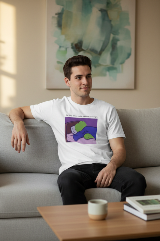 Man sitting on a couch wearing a White t-shirt with a graphic of a green pepe lying on a bed, with text 'Me trying to keep cool in the middle of the night' in a living room setting.