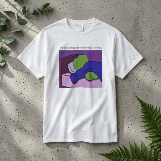 White t-shirt with a graphic of a green pepe lying on a bed, with text 'Me trying to keep cool in the middle of the night' on a textured surface with leaves.